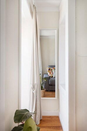A narrow hallway with a mirror reflecting a room with a gray sofa and a plant.