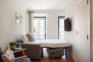 A compact room with a bed, a chair, a small table with a plant, and a TV mounted on the wall.