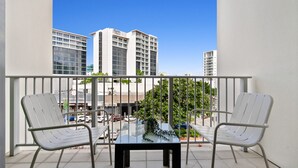 A balcony with two metal chairs and a glass table, overlooking a parking lot and buildings.