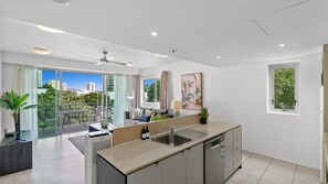 A modern kitchen with a large island, a dining area with a sofa and table, and a balcony with a view of the city. - Aveline Dual-Key at Mantra Trilogy | Pool & Gym Steps to Esplanade (Cairns City)