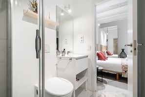 A modern bathroom with a white sink, mirror, and toilet. A bed with red pillows is visible in the adjacent room. - Dobohomes D14 (Madrid)