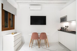 A modern kitchen with white cabinetry, a black flat-screen TV mounted on the wall, and a dining area with two pink chairs. - Dobohomes D14 (Madrid)