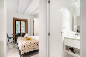 A bedroom with a bed, a bedside table with fruits, a chair, a window with a view, and a bathroom with a sink and mirror. - Dobohomes D14 (Madrid)