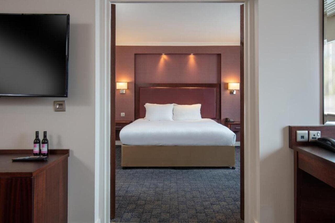 Ein Hotelzimmer mit einem großen Bett, einem flachbildfernseher an der Wand, einem Schreibtisch mit Telefon und zwei Wasserflaschen auf dem Schreibtisch.