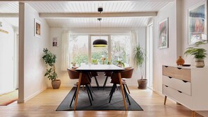 Un comedor con una mesa de madera, sillas y un tapete negro. Hay plantas en macetas y un mueble blanco con cajones.
