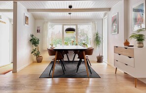 Un coin repas avec une table en bois, des chaises et un tapis noir. On y trouve également des plantes en pot et un meuble blanc avec des tiroirs.
