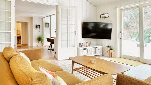 Una sala de estar con sofá, mesa de centro y televisor. Se ve una puerta que da a otra habitación y una ventana con vistas a árboles.