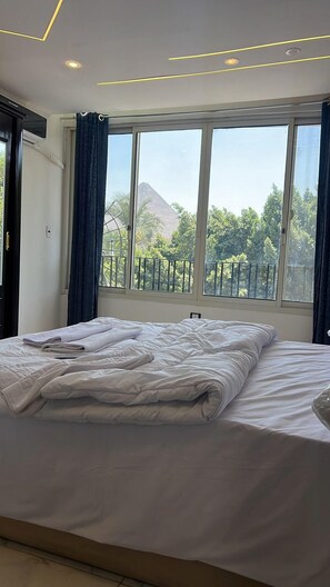 A modern bedroom with a large bed, a view of trees and a mountain through the window, and a balcony with a railing. - Miracle Pyramids Hotel (Giza)