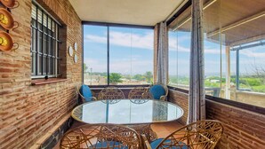 Un balcon avec une table en verre et des chaises en osier, des murs en briques et de grandes fenêtres offrant une vue sur la ville.