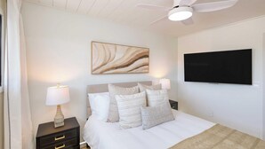 A bedroom with a bed, a ceiling fan, a mounted TV, and a bedside table with a lamp.