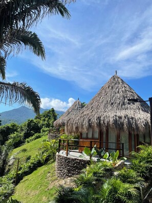 A thatched-roof building with a stone wall and a wooden deck, surrounded by lush greenery and a mountain in the background. - Zirwa Hotel Boutique (Santa Marta)