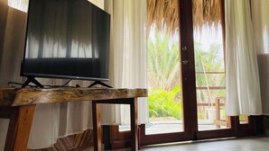 Ein Zimmer mit Fernseher auf einem Holzboden, weißen Vorhängen, einem Reetdach und Blick ins Grüne.