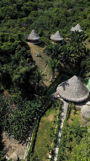 Luchtbeeld van een resort met rieten hutten, omringd door weelderige tropische begroeiing en een helder waterpartij.