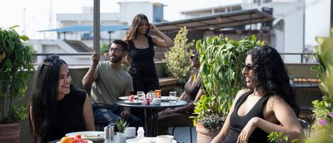 Un grupo de personas sentadas en mesas al aire libre, con plantas en macetas y un edificio de fondo.