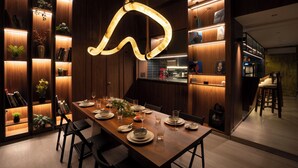 A modern dining area with a wooden table set for a meal, surrounded by bookshelves and a kitchen area. - Hotel PXL 27 (Bengaluru)