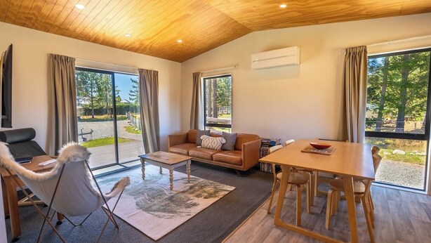 Ein modernes Wohnzimmer mit Holzbalkendecke, einem Ledersofa, einem Esstisch und großen Fenstern mit Blick ins Freie.