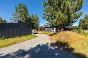 A modern house with a gravel driveway, surrounded by tall trees and a metal fence. - North West Cabin - private high country escape. (Twizel)