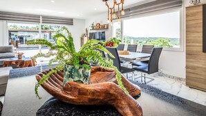 Un salon moderne doté d’une grande fenêtre, un coin repas avec une table et des chaises, et une composition végétale originale présentée dans un récipient en bois aux formes travaillées.