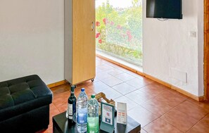 Une pièce avec un canapé noir, une petite table sur laquelle sont posées des bouteilles d’eau, un téléviseur fixé au mur et une vue sur la verdure par une grande fenêtre.