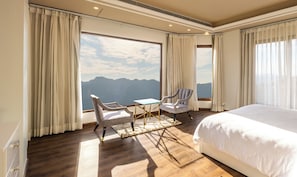Une chambre d’hôtel avec un grand lit, deux fauteuils, une petite table et une vue sur les montagnes grâce à de grandes fenêtres.