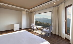 A bedroom with a large bed, a desk, two chairs, a dresser, and a view of mountains.