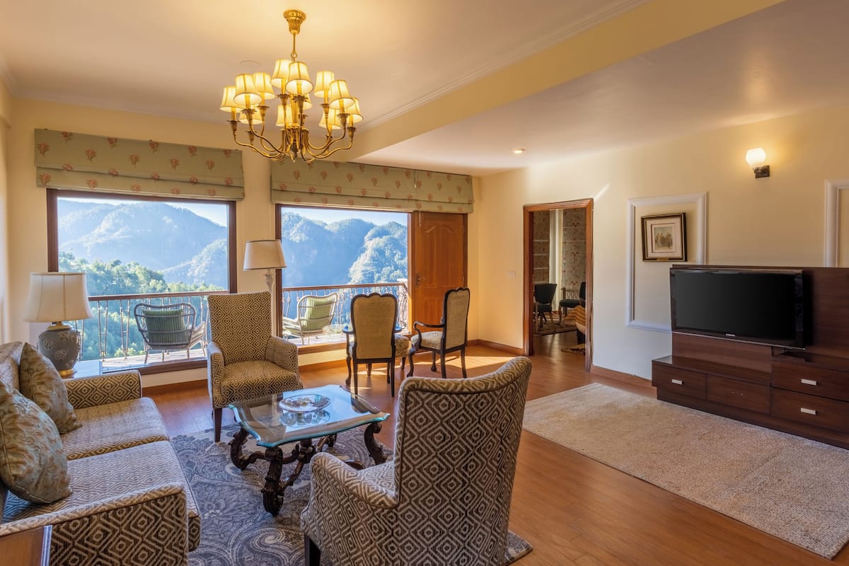 Ein Wohnzimmer mit einer Couch, Sesseln, einem Couchtisch, einem Fernseher und einem Balkon mit Bergblick.