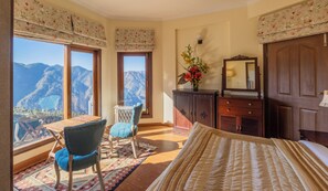 A bedroom with a large window offering a mountain view, a wooden dresser, a chair, and a bed with a patterned bedspread.