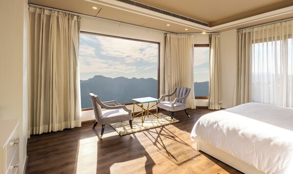 A hotel room with a large bed, two chairs, a small table, and a view of mountains through large windows.