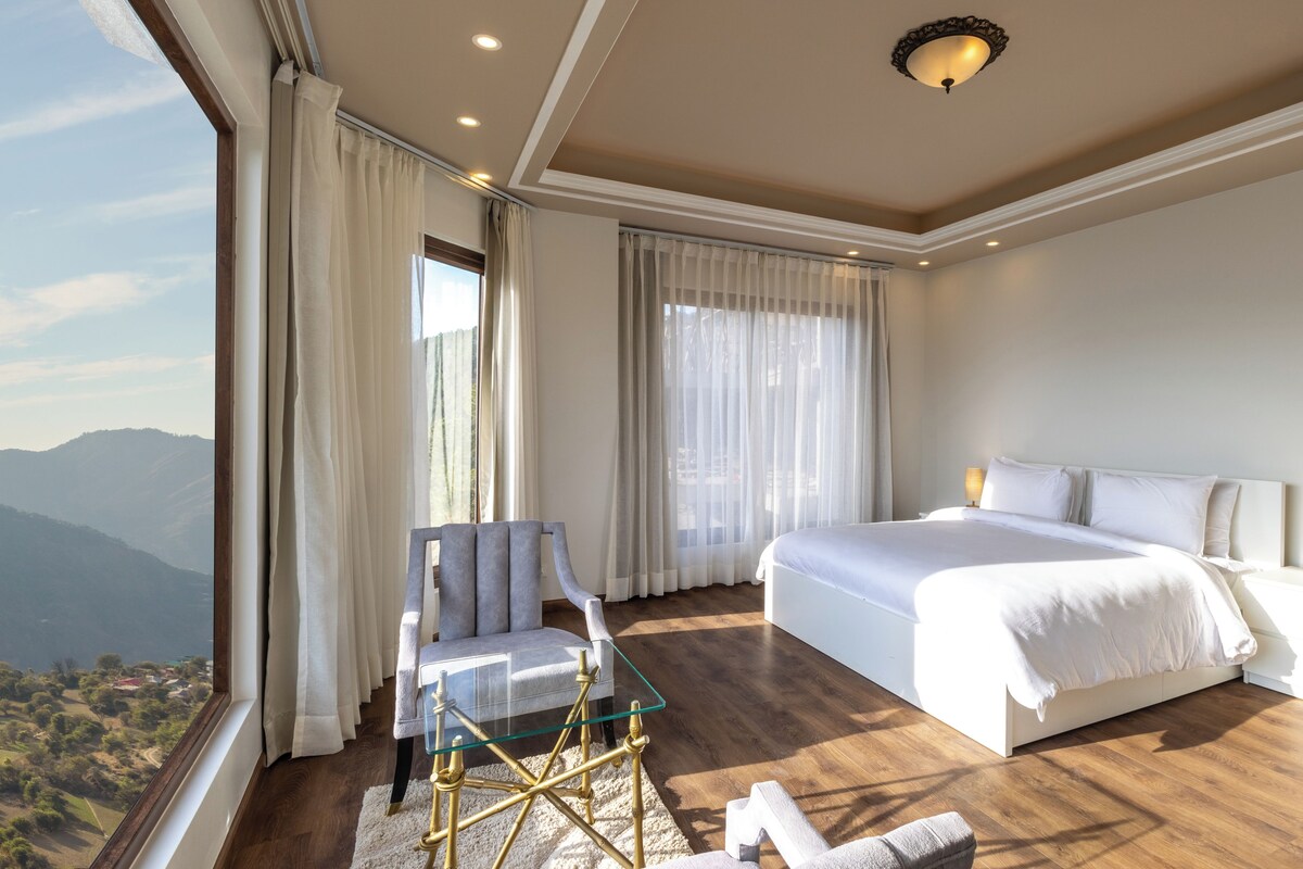 A bedroom with a bed, a chair, a glass table, and a view of the mountains.