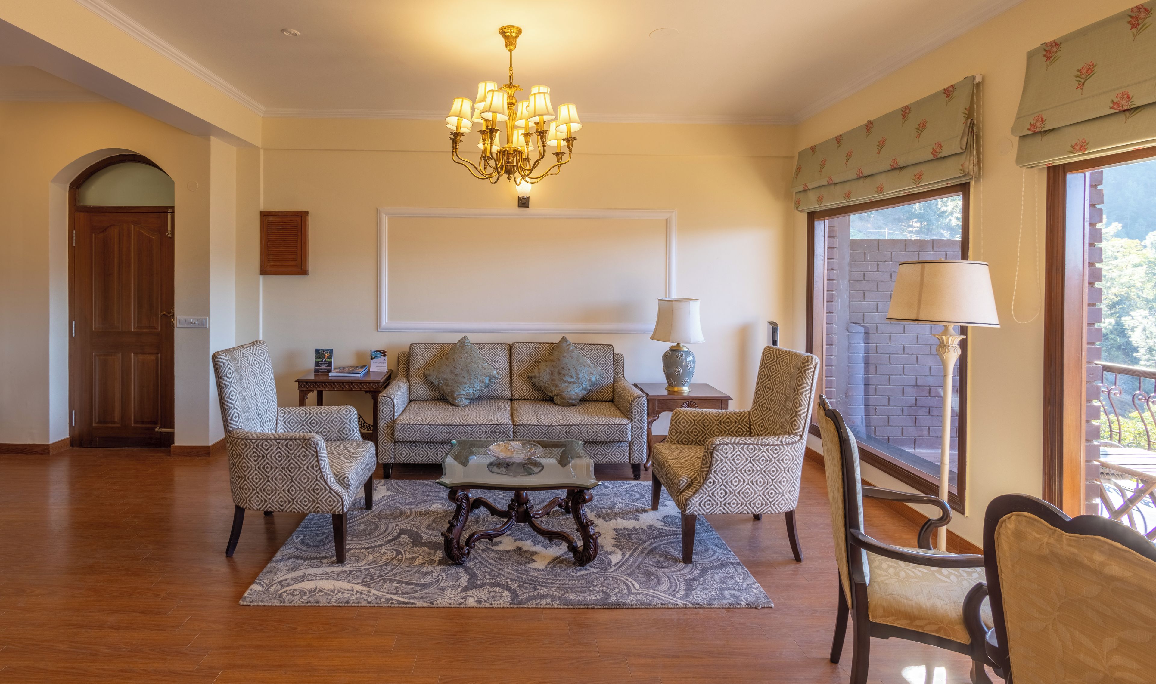 A living room with a sofa set, patterned chairs, a coffee table, a chandelier, and a view through a window.