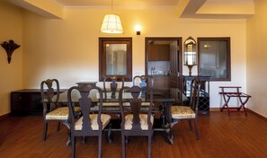 A dining area with a glass table, wooden chairs, a chandelier, and a kitchen in the background.