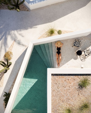 A rooftop pool with a clear blue water feature, surrounded by a white pool deck and sparse desert vegetation. - Anome Hotel (Mykonos)