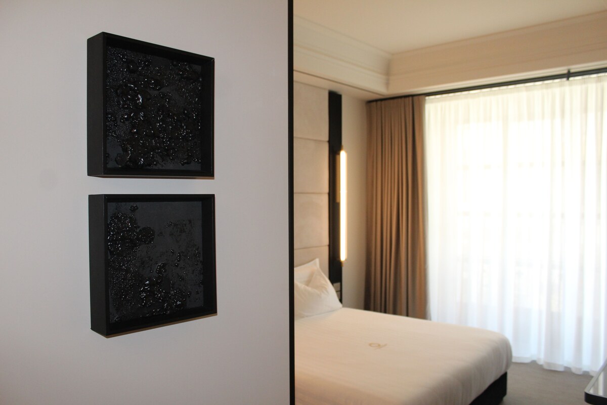 Una habitación de hotel con una cama, dos cuadros enmarcados en la pared y un ventanal con cortinas.