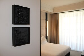 Una habitación de hotel con una cama, dos cuadros enmarcados en la pared y un ventanal con cortinas.