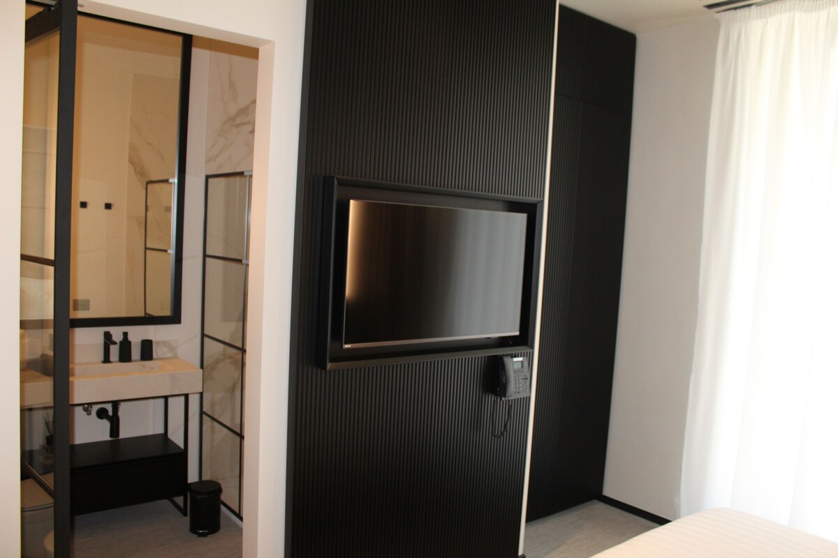 Habitación de hotel moderna con un televisor de pantalla plana grande montado en un panel negro, baño con espejo, lavamanos y retrete, y una ventana grande con cortinas.