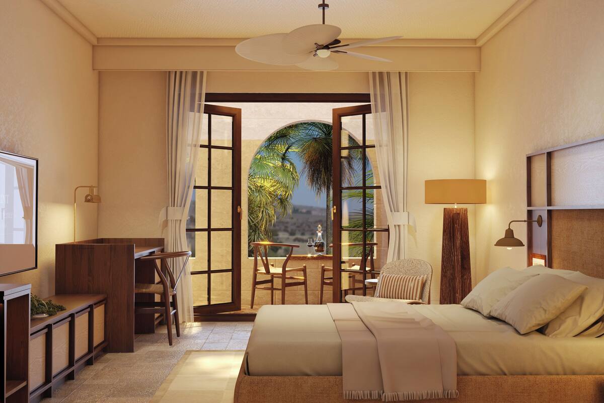 Ein Hotelzimmer mit Bett, Schreibtisch, Stuhl und Blick auf Palmen durch eine offene Tür.