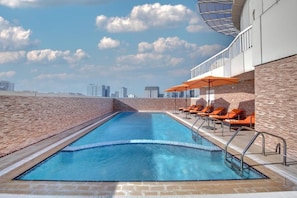 A rooftop swimming pool with a city skyline in the background, surrounded by brick walls and orange lounge chairs. - City Stay Hotel Expo Center Sharjah (Sharjah)