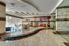A modern hotel lobby with a curved marble reception desk, seating area with tables and chairs, and large windows offering views of the street. - City Stay Hotel Expo Center Sharjah (Sharjah)
