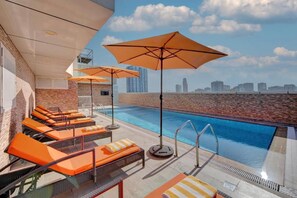 Ein Poolbereich auf dem Dach mit orangefarbenen Liegestühlen und Sonnenschirmen, im Hintergrund eine klare Aussicht auf die Stadtlandschaft.