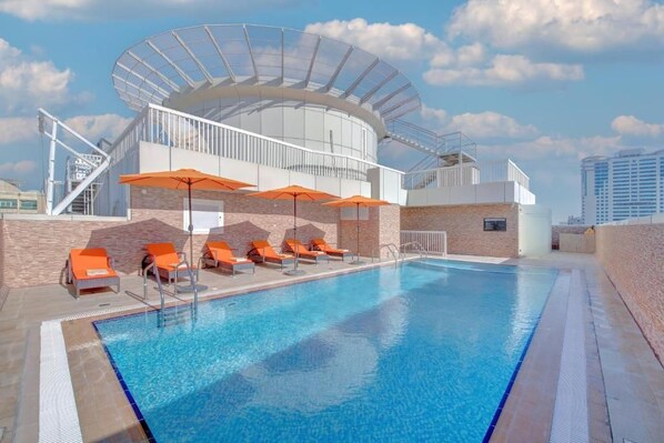 A swimming pool with lounge chairs and umbrellas, a covered seating area, and a modern building with a glass facade. - City Stay Hotel Expo Center Sharjah (Sharjah)