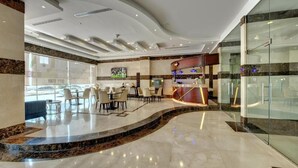 A modern hotel lobby with a curved marble reception desk, seating area with tables and chairs, and large windows offering views of the street. - City Stay Hotel Expo Center Sharjah (Sharjah)