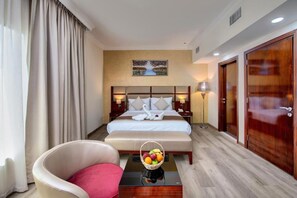 A hotel room with a large bed, a chair, a bedside table with a fruit basket, and a painting on the wall. - City Stay Hotel Expo Center Sharjah (Sharjah)