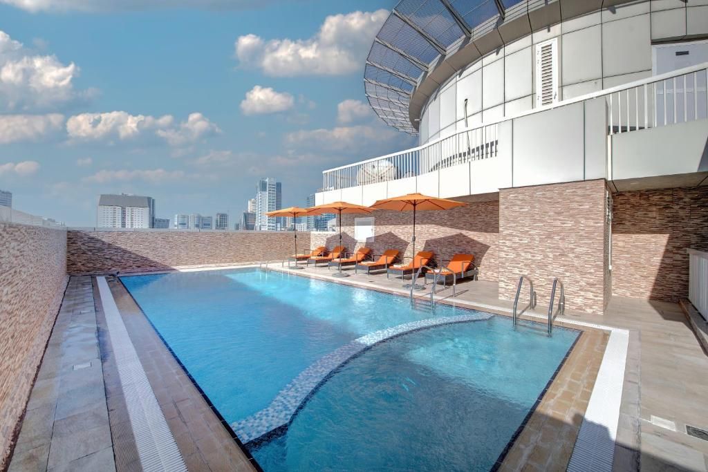 A rooftop swimming pool with lounge chairs and umbrellas, surrounded by a modern building with a glass facade.