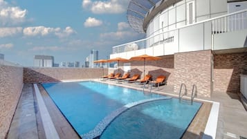 A rooftop swimming pool with lounge chairs and umbrellas, surrounded by a modern building with a glass facade.