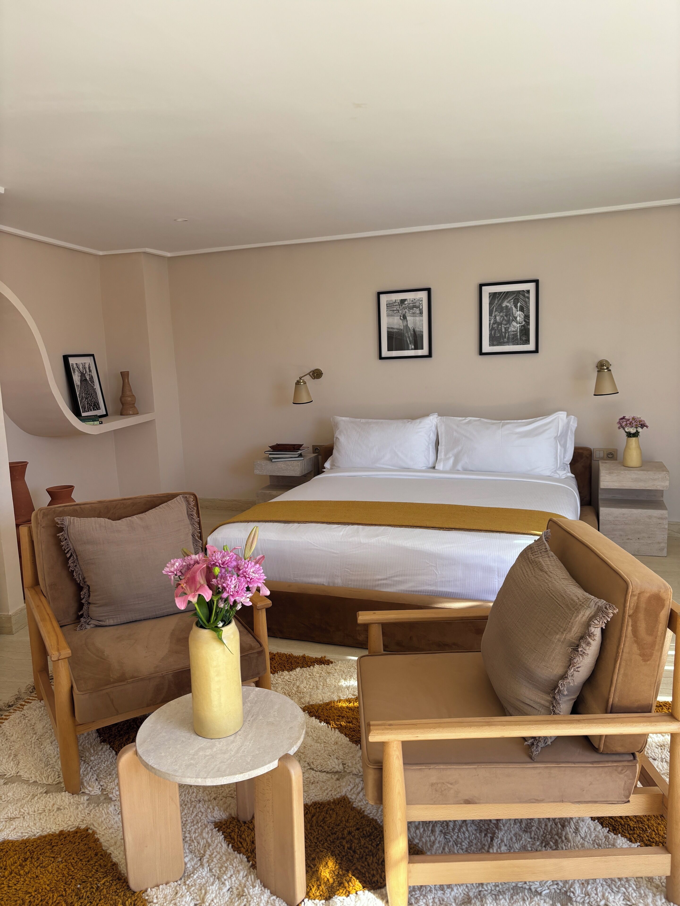 A bedroom with a bed, two chairs, a small table, and a nightstand with a lamp.