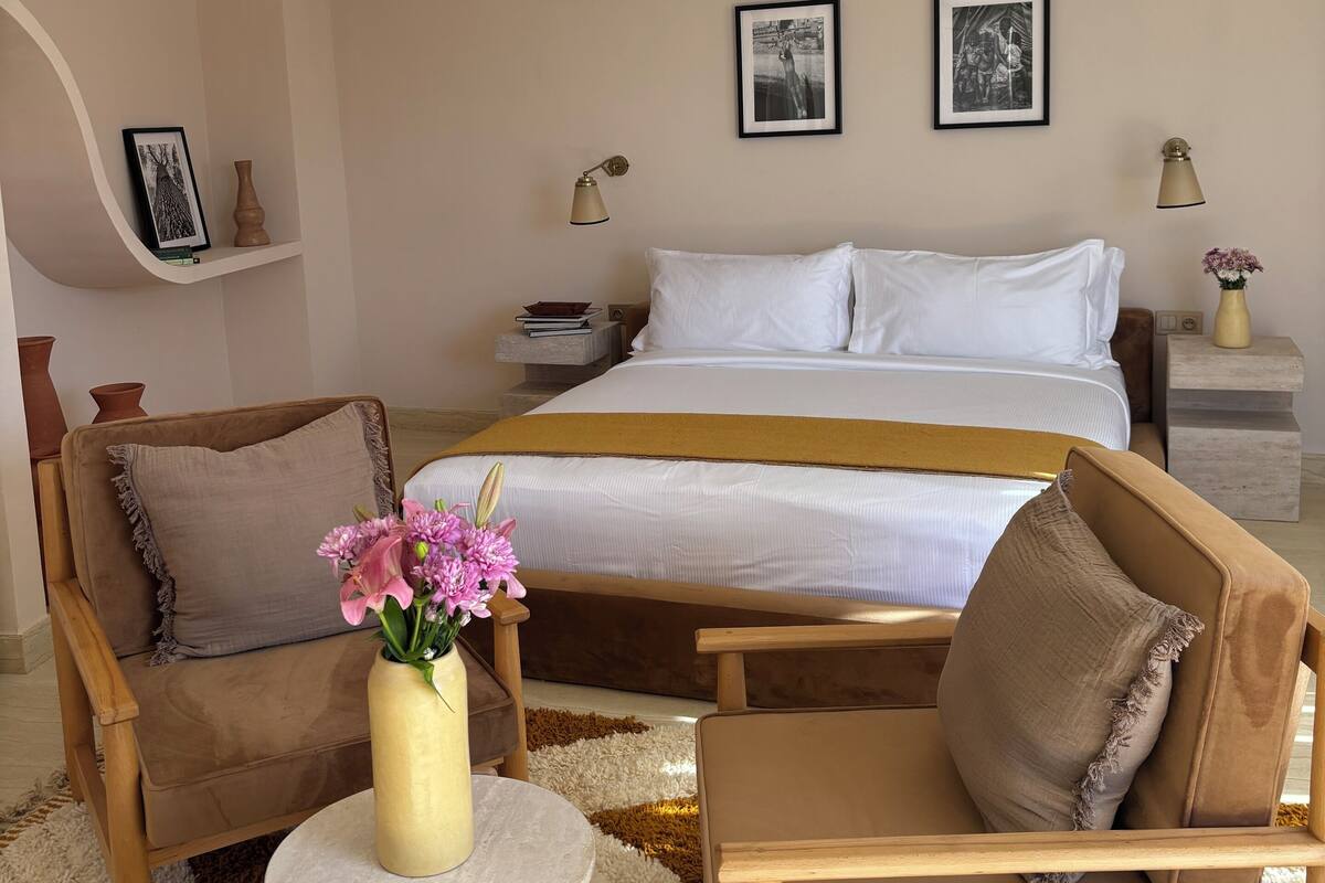 A bedroom with a bed, two chairs, a small table, and a nightstand with a lamp.