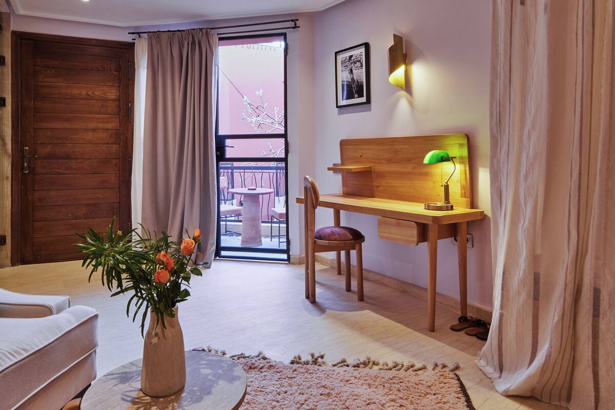A modern living room with a wooden desk, a small table with a vase, a sofa, and a view of the outside balcony.