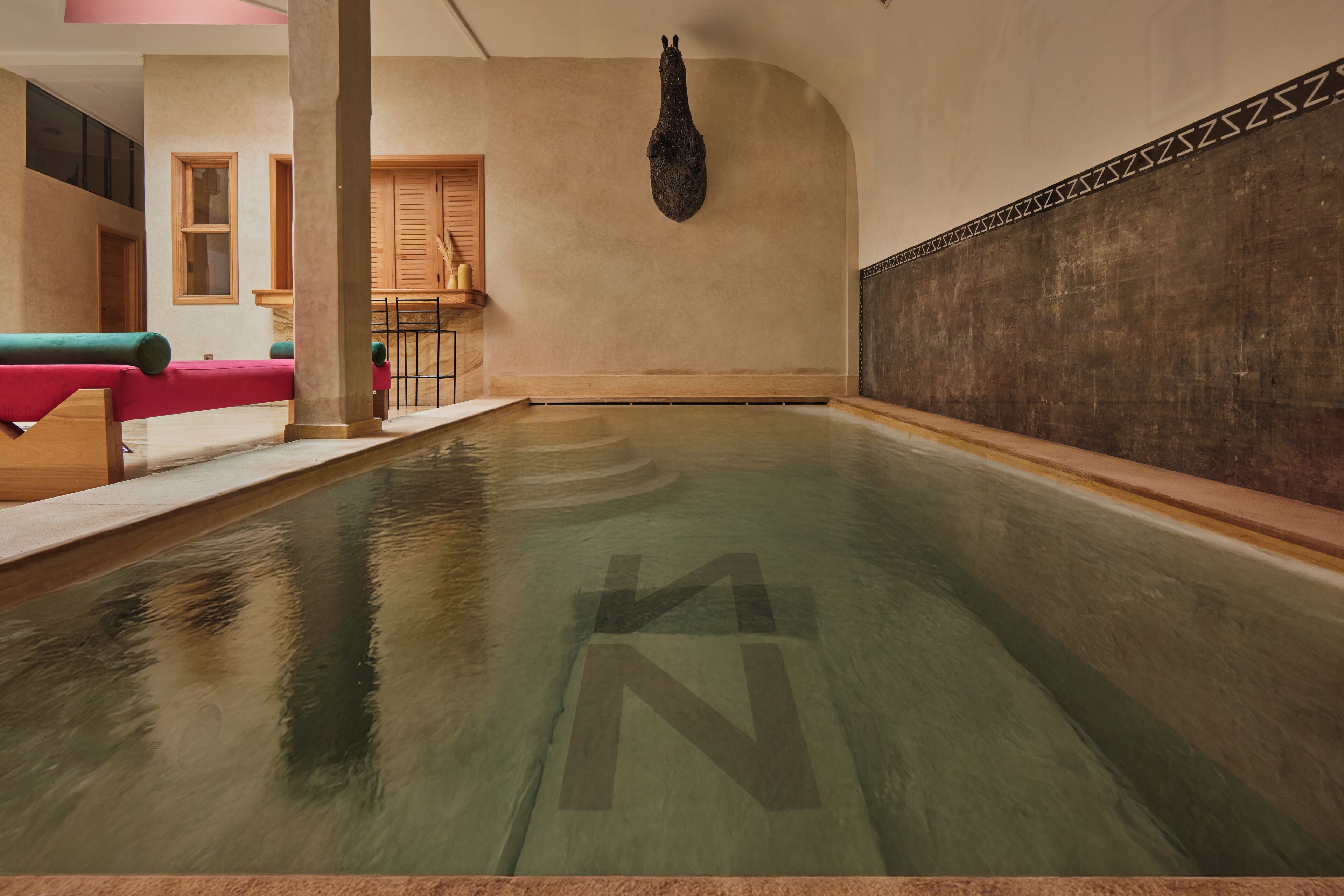 A large indoor pool with a clear 'N' symbol on the floor, wooden benches, and a stone wall with a decorative pattern.