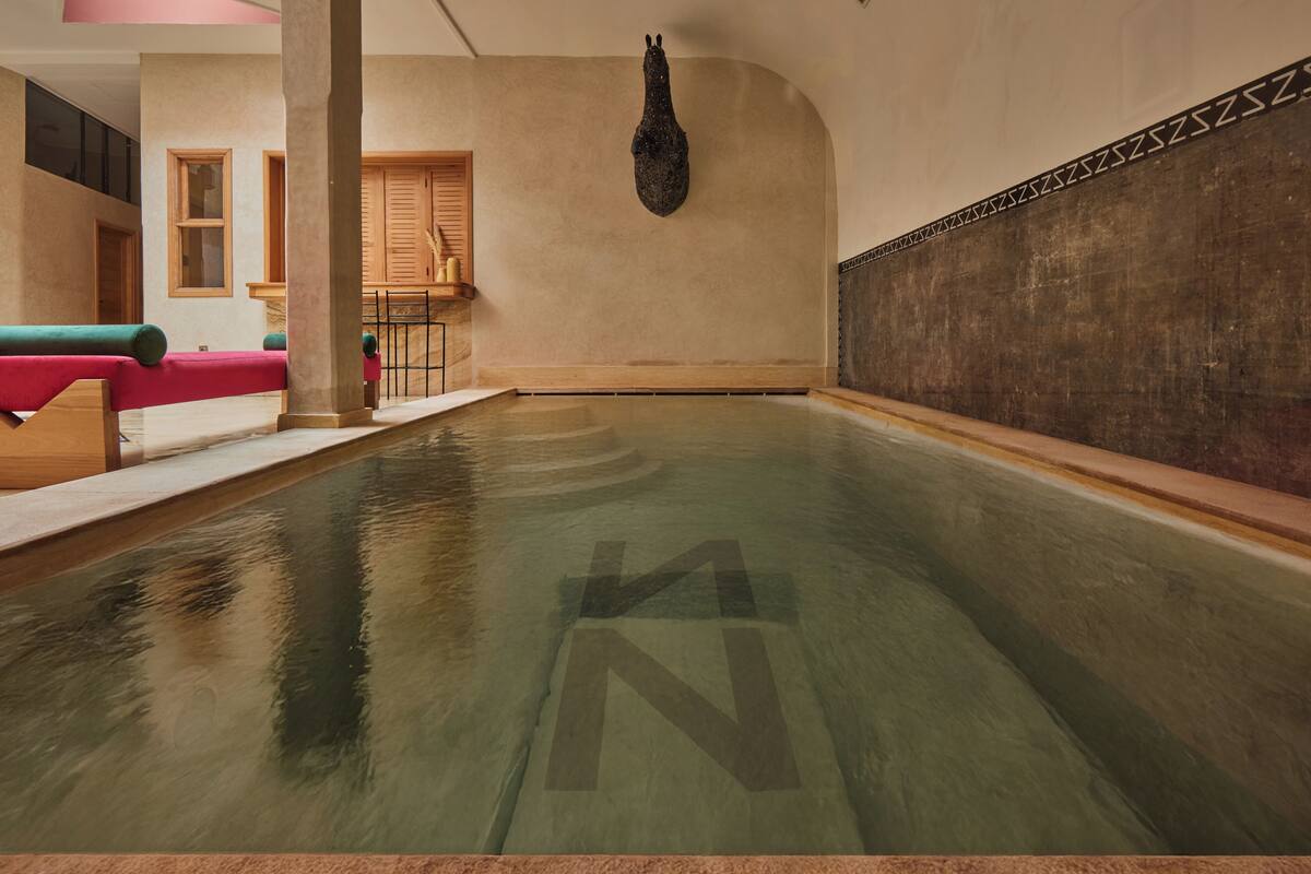 A large indoor pool with a clear 'N' symbol on the floor, wooden benches, and a stone wall with a decorative pattern.