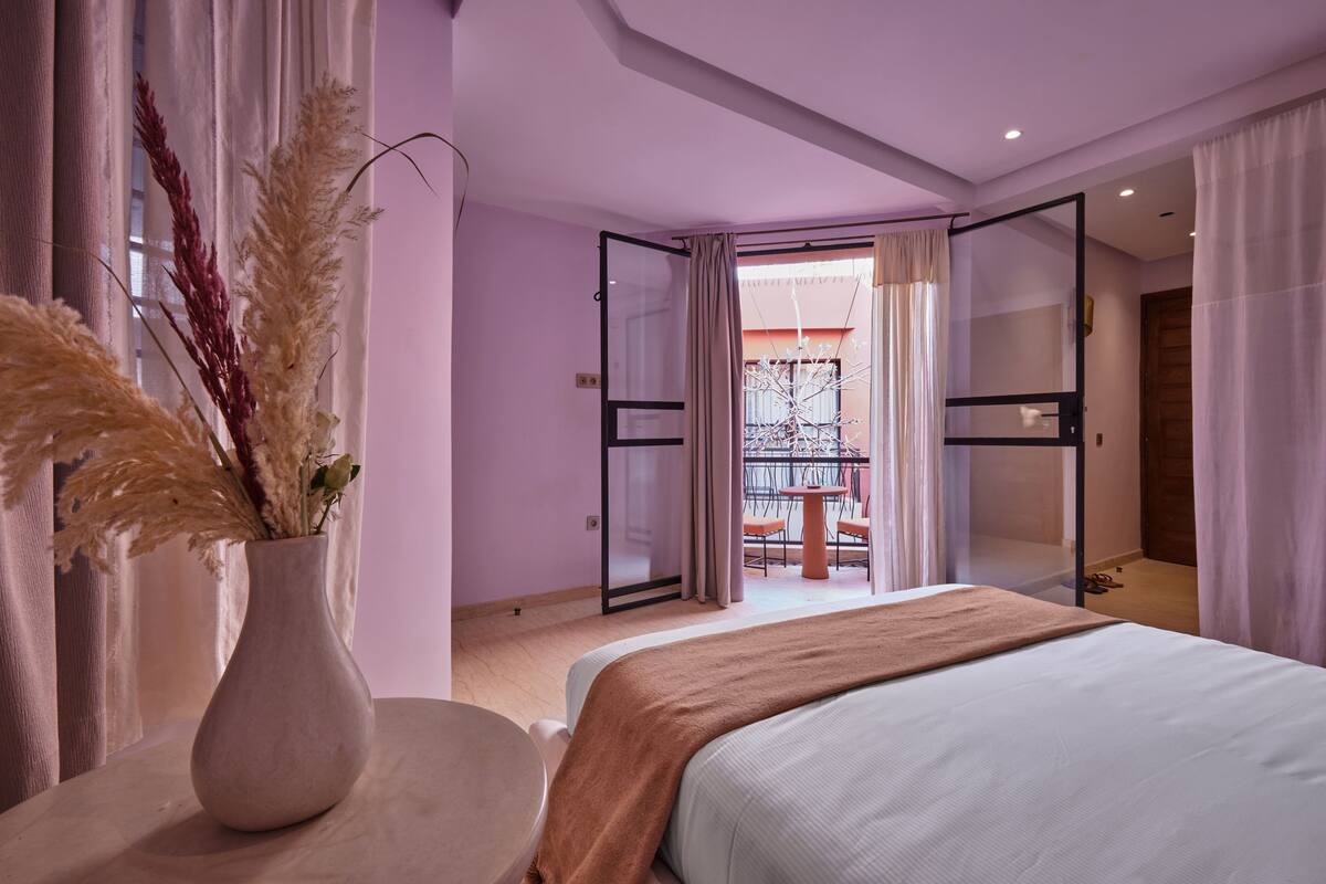 A bedroom with a bed, a vase with dried plants, a table, and a balcony with a railing.